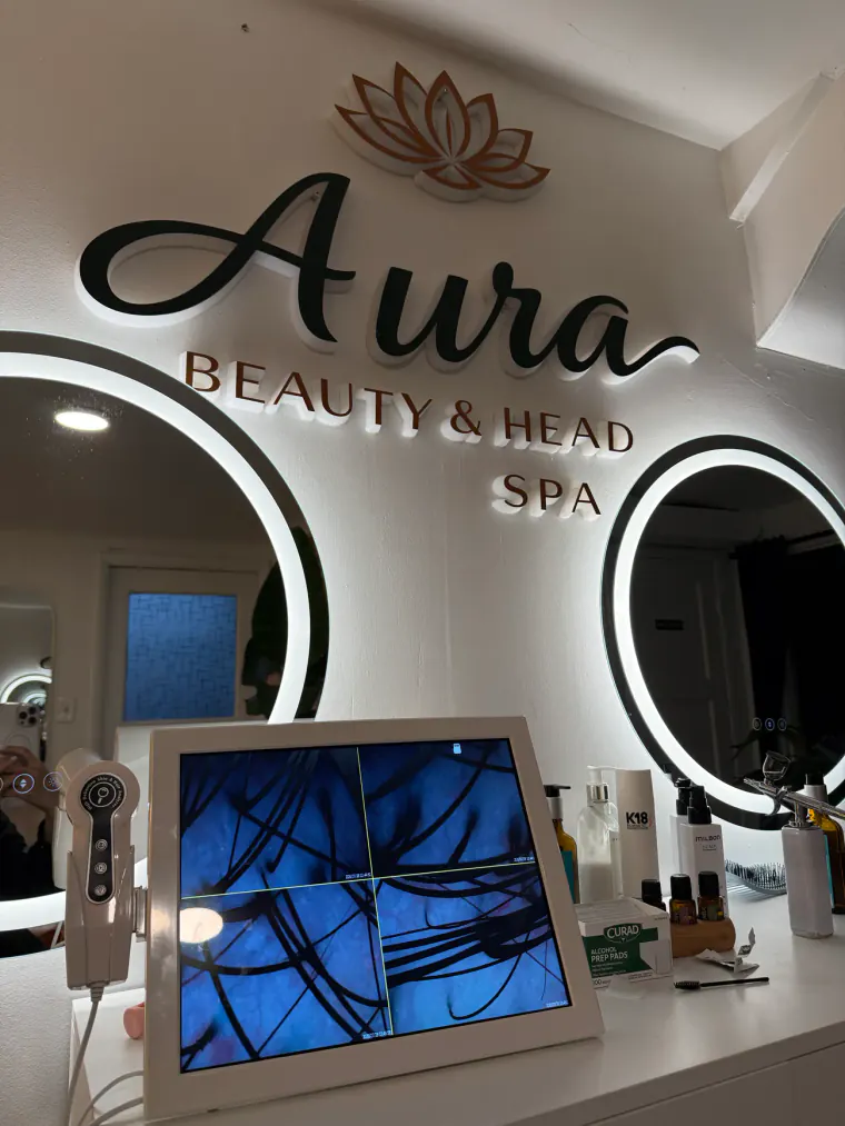 The handheld scalp camera at Aura in Issaquah beside a monitor showing four magnified views of a scalp, under the backlit Aura wordmark